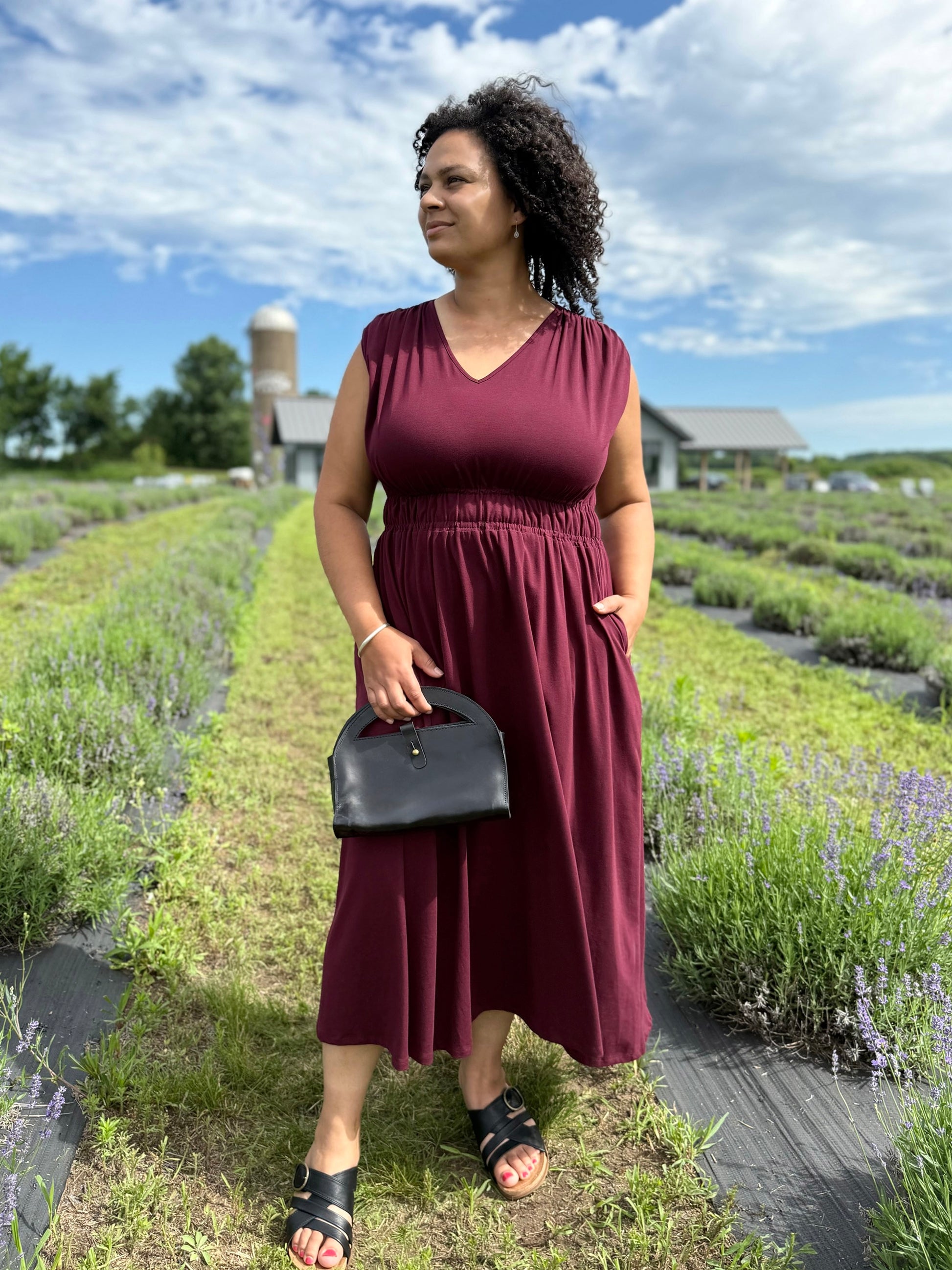 Woman in a burgundy dress standing in a lavender field with a black leather handbag.