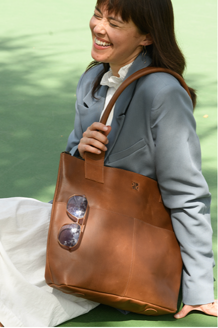 Woman holding a brown leather tote bag with sunglasses on a green outdoor background
