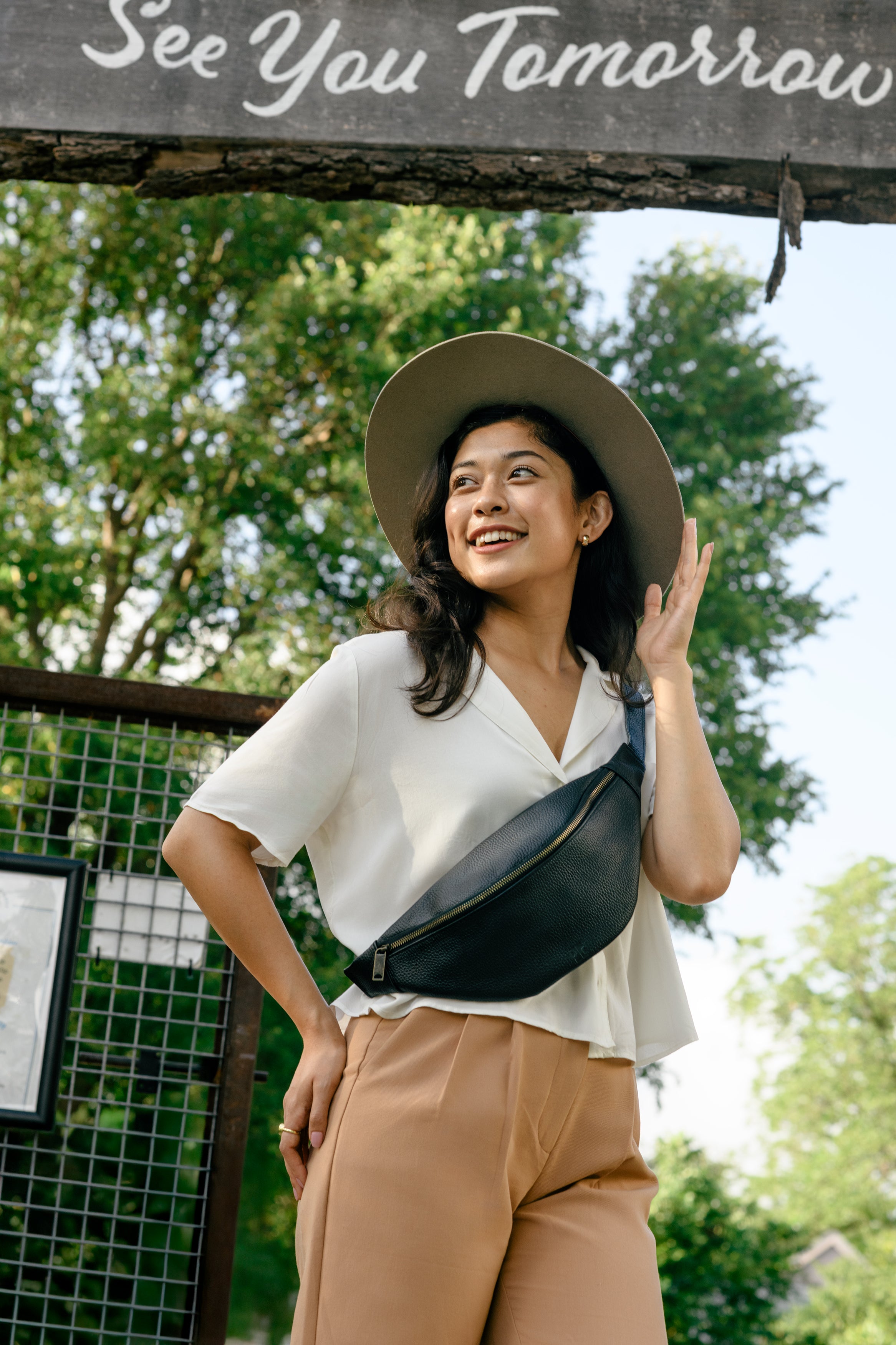 Woman wearing a white shirt, beige pants, and a wide-brimmed hat with a black fanny pack, standing outdoors with 'See You Tomorrow' sign above.