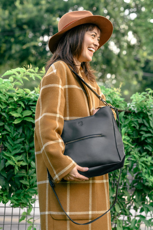 Woman in a plaid coat and hat holding a black leather handbag outdoors.